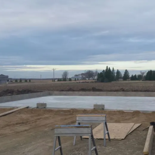 Bare outdoor construction site with concrete foundation, dirt lot, and cloudy sky beyond a rural view