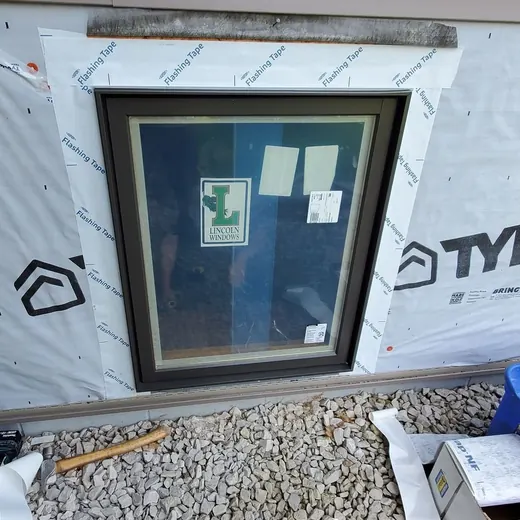 Basement window well under white siding, bordered by gravel and insulation scraps