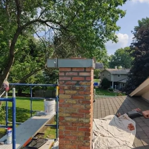 Brick chimney under construction on a rooftop, with a tree and suburban homes in the background.