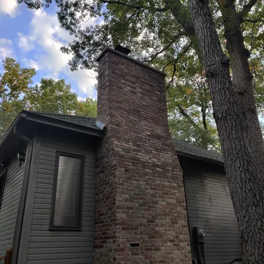 Brick chimney on a dark house, framed by tall trees and a cloudy blue sky.