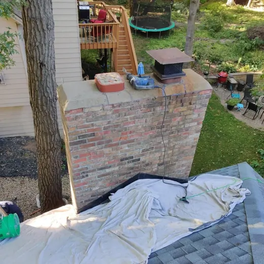 Brick patio wall with potted plants and a trampoline in a leafy backyard; tarp spread across shingles in foreground