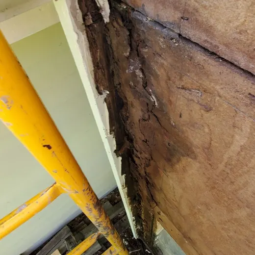 Close-up of a damaged wall seam with peeling material, exposed wood, and construction equipment below