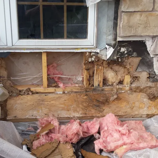 Damaged brick wall beneath a window, with insulation and debris exposed in the lower section.