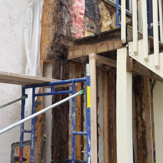 Damaged exterior wall with exposed insulation, scaffolding, and a white balcony railing above.