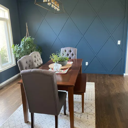 Dining room with dark blue walls, wooden table, tufted chairs, and rug by a bright window.