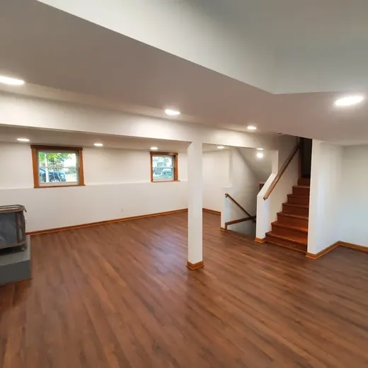 Empty basement living area with wood floor, white walls, fireplace, windows, and stairs to the upper level