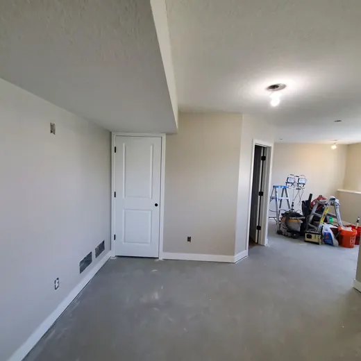 Empty carpeted room with white walls, closet doors, and a hallway opening to cluttered boxes and tools.