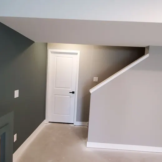 Empty gray carpeted room with white trim, a doorway, and stairs along the right wall.