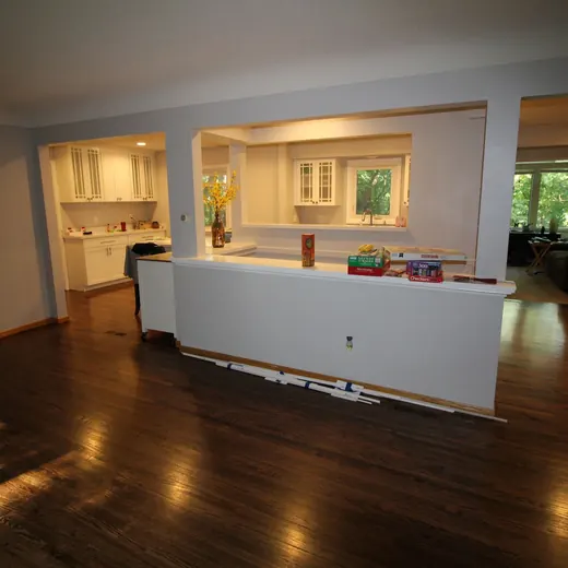 Empty living room with dark wood floors, white half wall, and a bright adjoining kitchen area.