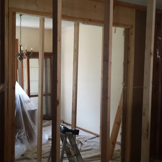 Exposed wooden studs in an unfinished room with construction tools and a ladder near a window.