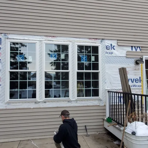 Exterior of a house under construction with new windows, siding wrap, and a person on the deck.