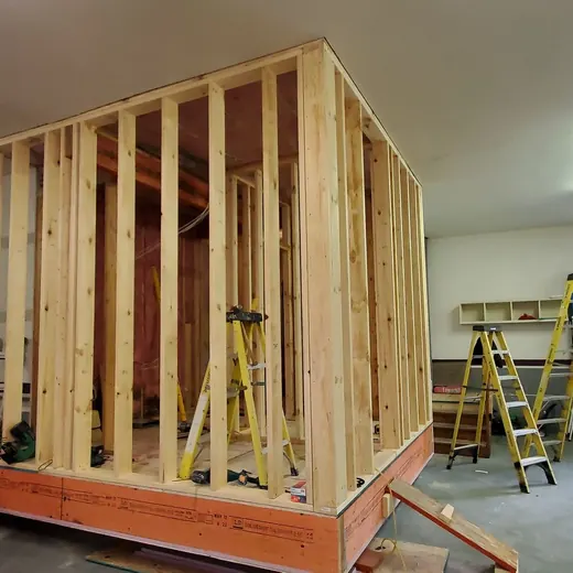 Framed wooden room under construction in a garage, with ladders, tools, and a workbench nearby