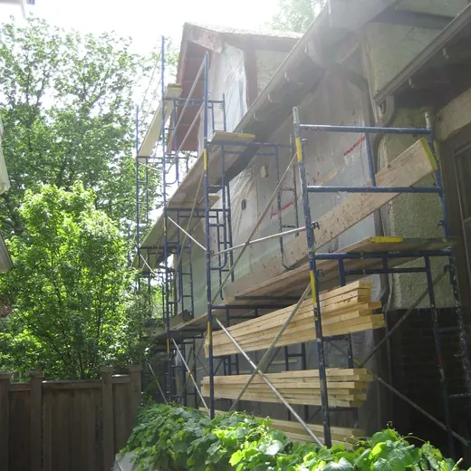 House under construction with blue scaffolding beside a leafy backyard fence