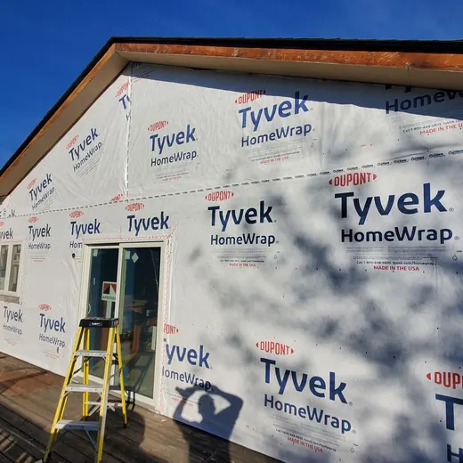 House under construction wrapped in Tyvek, with a ladder and blue sky visible.