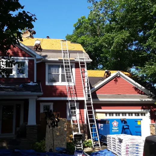 House with workers on roof and ladder, red siding, blue tarp, and truck in driveway