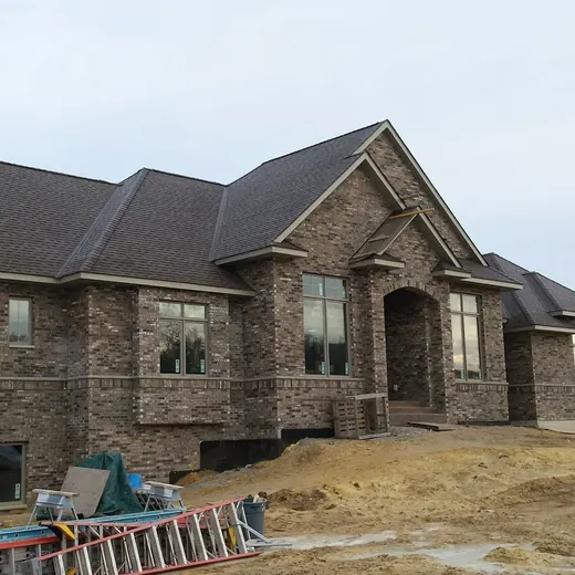 Large stone house under construction on a dirt lot, with roof shingles and construction materials nearby