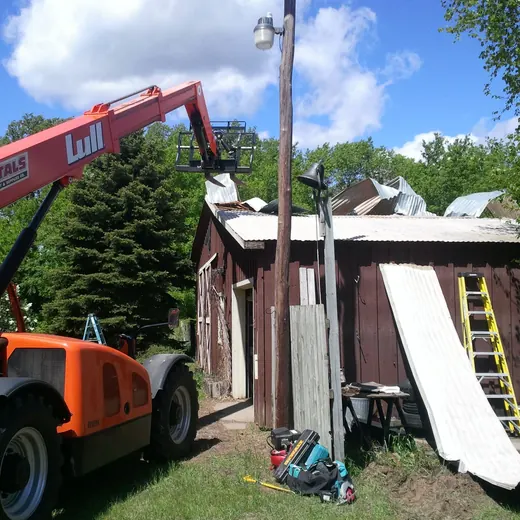 Orange excavator lifting debris from a damaged shed, with a yellow ladder nearby.