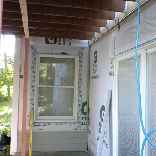 Partially built porch with exposed wood ceiling, window, sliding door, and construction wrap on the walls.