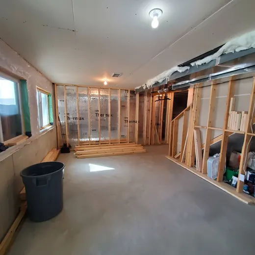 Partially renovated room with exposed studs, windows, and a trash bin on a concrete floor