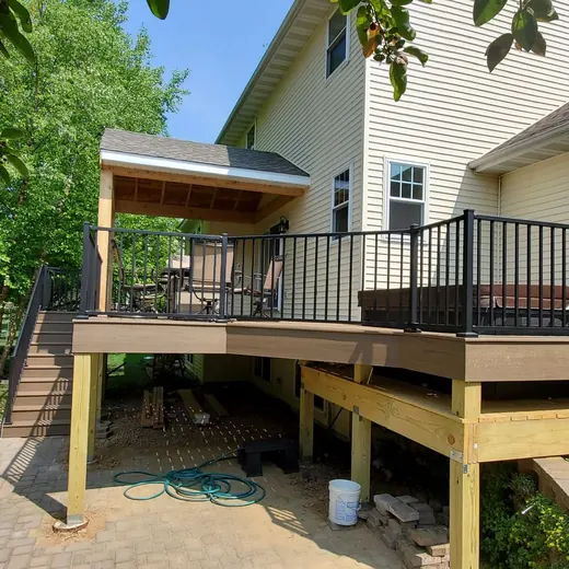 Raised wooden deck with black railing beside a white house, overlooking a stone patio and green yard