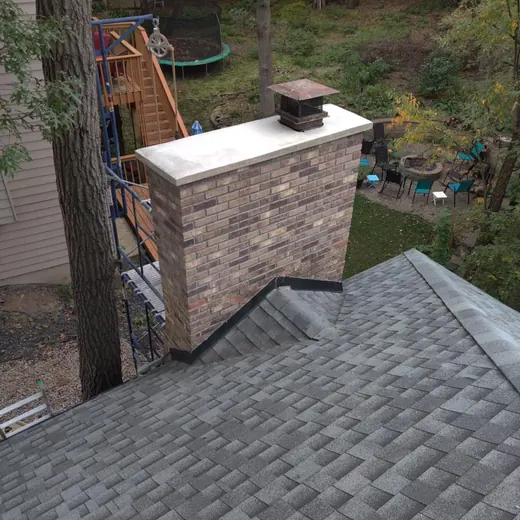 Rooftop view of a brick chimney beside gray shingles, with a wooded backyard and playground in the background