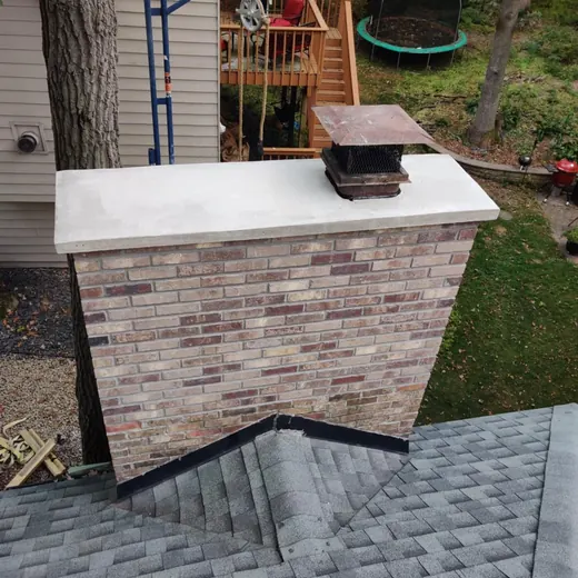 Rooftop chimney with brick surround and metal cap, overlooking a backyard with trees and a trampoline.