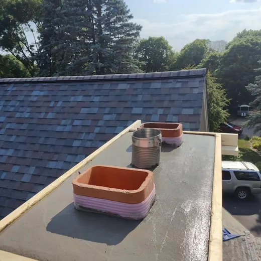 Rooftop with freshly coated black flat roof and two orange vent chimneys beside blue-shingled roof.