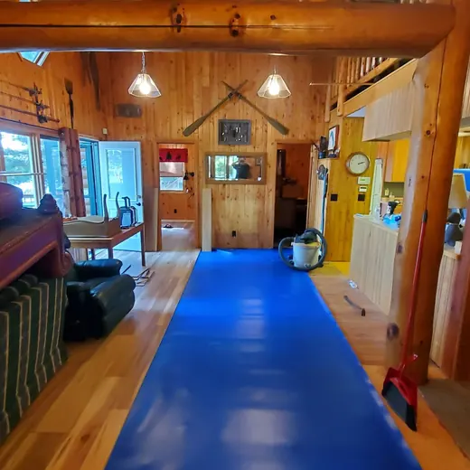 Rustic cabin interior with wooden walls, a blue runner, and a dining area visible through the doorway