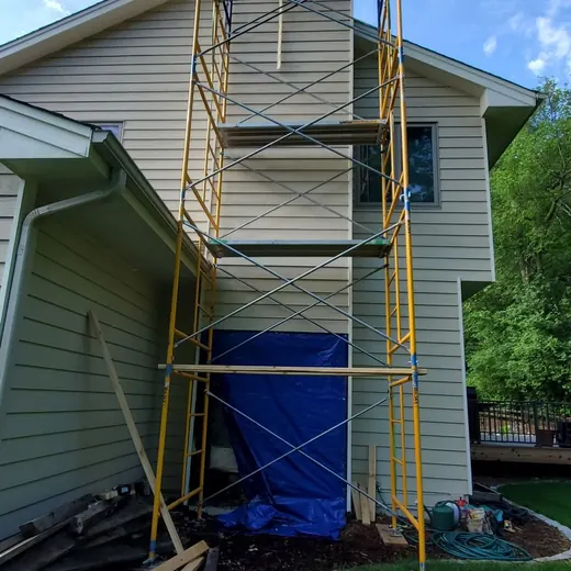 Scaffolding around a house exterior with a blue tarp at the base and ladder frames above