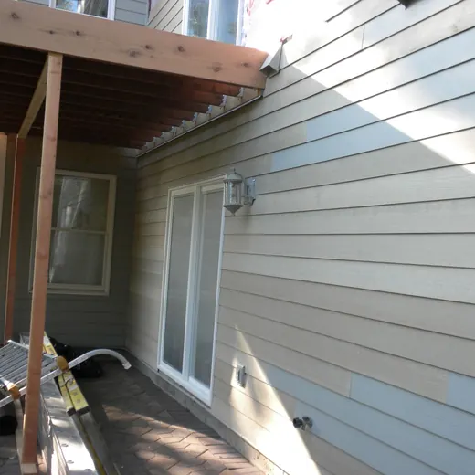 Side of a gray house with a covered porch, windows, and construction materials along the wall.
