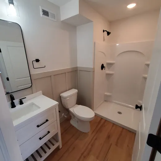 Small bathroom with white vanity, toilet, wood floor, and walk-in shower with built-in shelves.