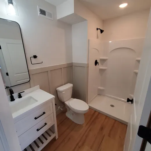 Small white bathroom with wood floor, vanity, toilet, and walk-in shower.