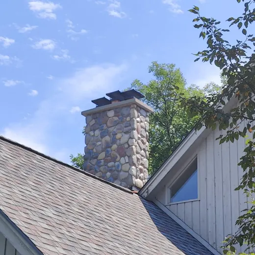 Stone chimney on a house with a shingled roof under a blue sky with clouds