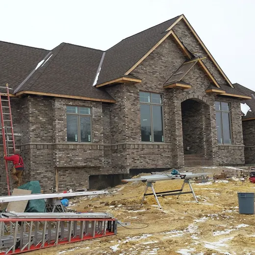 Stone house under construction with roof framing, ladders, and snowy ground