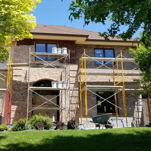 Two-story house under renovation with scaffolding and white tarp, surrounded by green trees and lawn