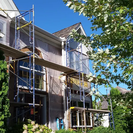 Two-story house with scaffolding and ladder under construction, viewed beside a tree on a sunny day