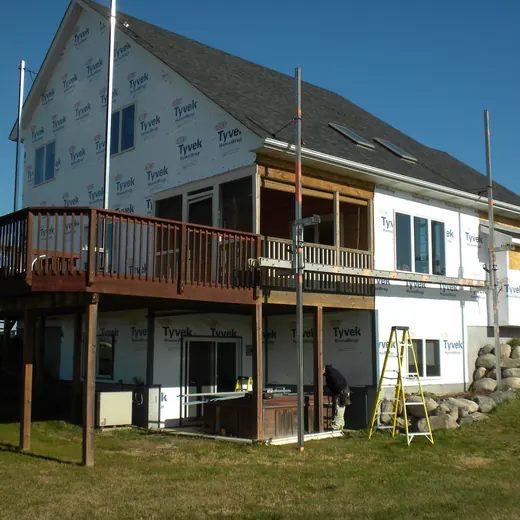 Two-story white house with a wooden deck and balcony on a grassy hillside under blue sky