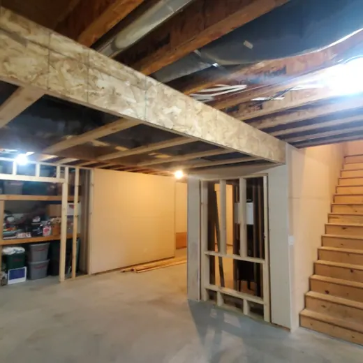 Unfinished basement with exposed beams, concrete floor, storage area, and wooden stairs leading up