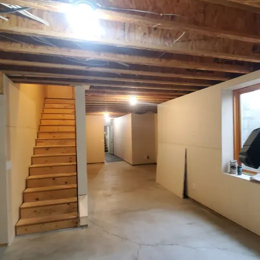 Unfinished basement with exposed beams, stairs, hallway, and a window on the right.
