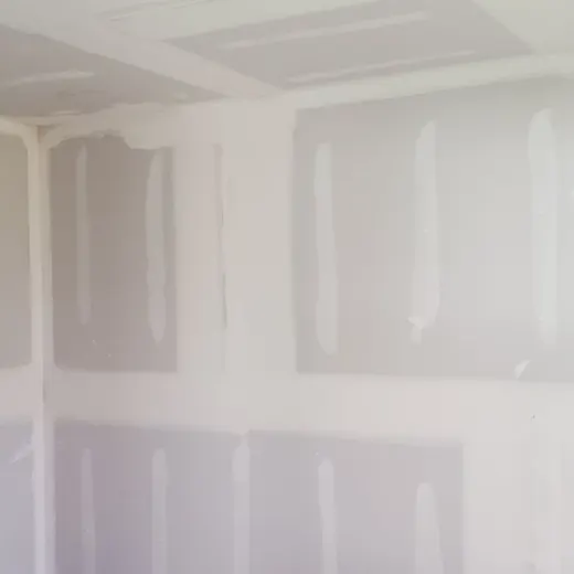 Unfinished drywall in a room, with taped seams and patches on white walls and ceiling.