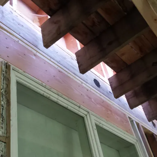 Upward view of house eaves with exposed rafters above a window and beige siding