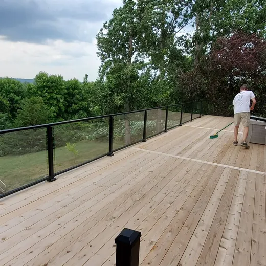 Wide wooden deck overlooking green hills, with two people near a house and outdoor seating.
