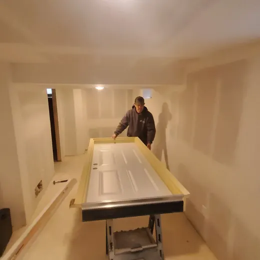 Worker in a narrow unfinished room moving a large white panel on a cart during renovation.