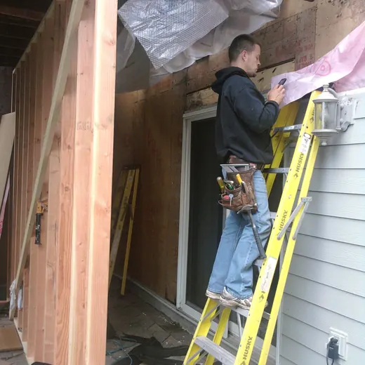 Worker on yellow ladder repairing exterior wall and soffit under a porch overhang.