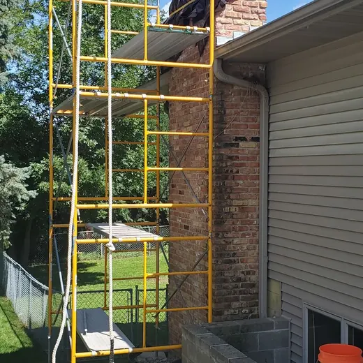 Worker on yellow scaffold outside a brick house beside a white siding wall under a blue sky