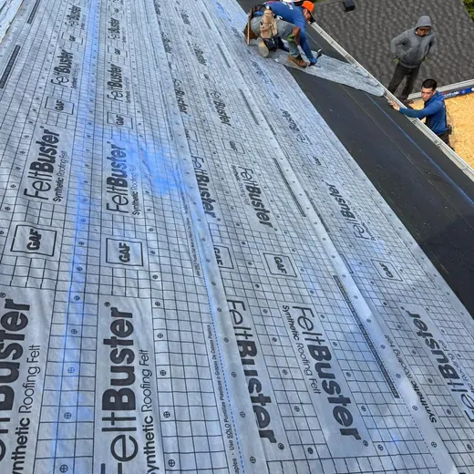 Workers installing roofing underlayment on a sloped roof, with blue and gray synthetic sheets visible.