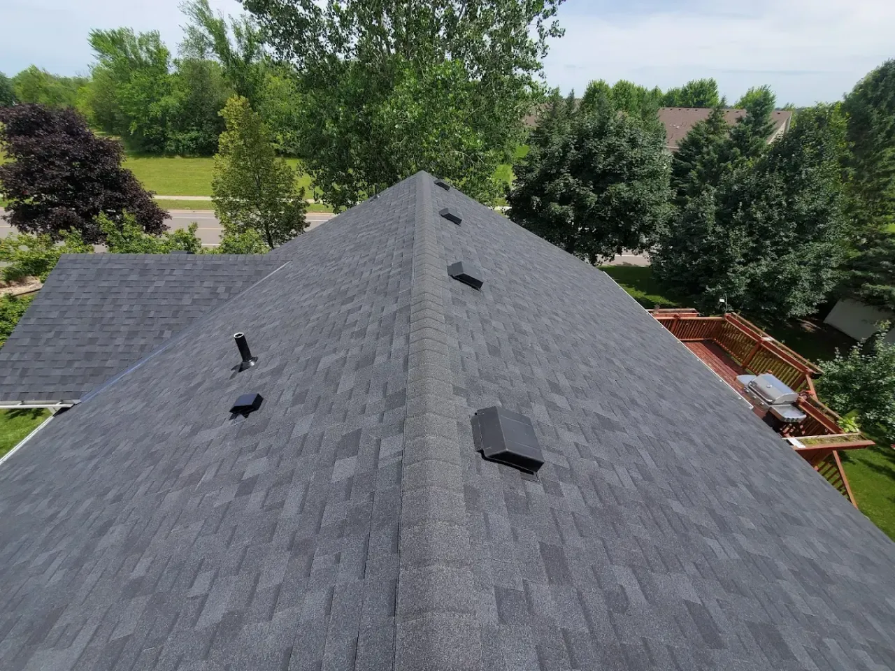 Dark gray shingled roof with vents, viewed from above against trees and neighboring houses