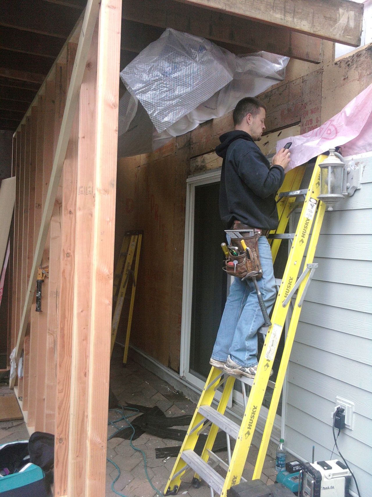 Worker on a yellow ladder repairing the exterior of a wooden house under a porch roof.