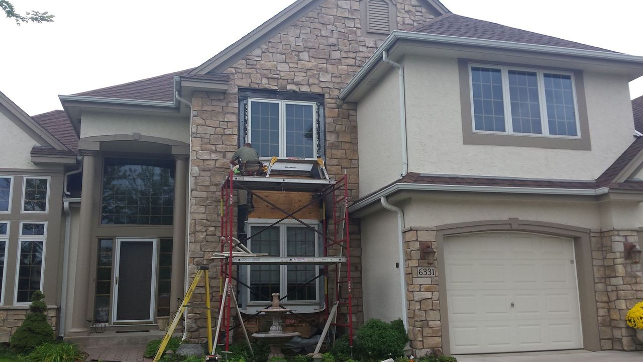 Two-story suburban house with stone facade, garage, and ladders/scaffolding in front, suggesting exterior work