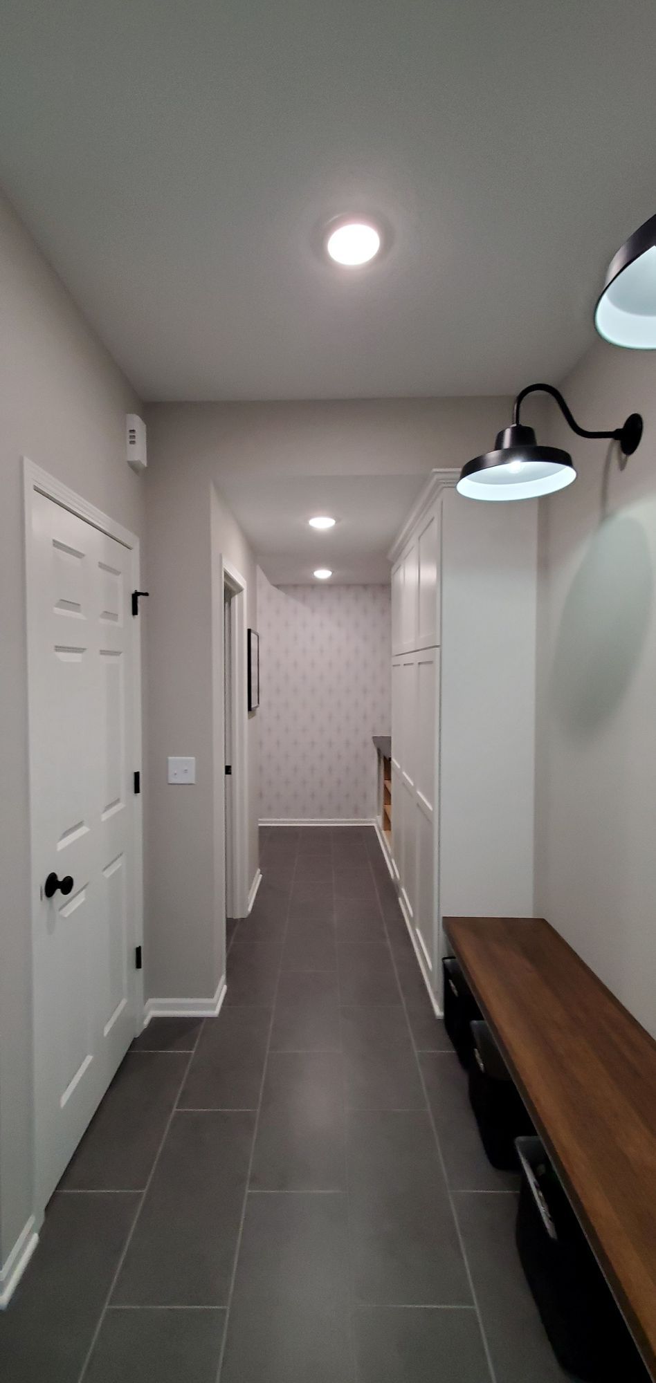 Bright hallway with gray tile floor, white walls, doors, wall lights, and a wooden bench.